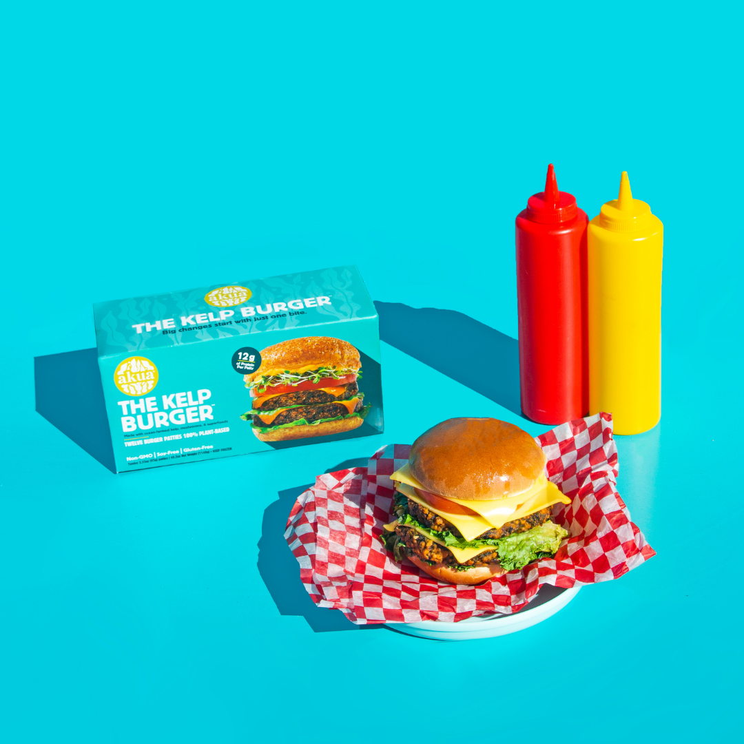 Stacked Kelp burger with multiple patties, cheese, veg and buns, next to its box, mustard and ketchup