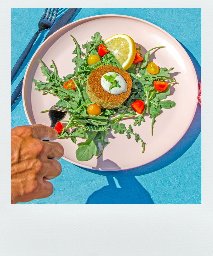 Poloroid-style photo of Kelp Krab Cake on a plate on a bed of rocket, tomatoes and garnished with sauce and a lemon. A hand with fork is reaching in to take some